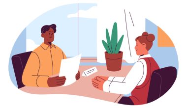 Two people seated at a table in an office, one presenting a paper while the other listens, with a potted plant and window in the background.