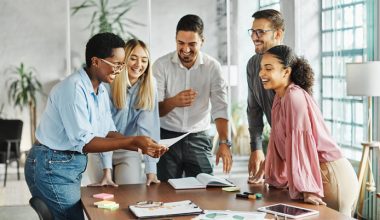 A group of young workers stand around a table while one person holds a piece of paper for everyone to look at. There are two men and three women of different ethnicities.