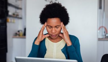 A woman is sitting at a table in a kitchen with an open laptop in front of her and her head in her hands.