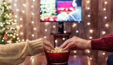 A close up photo of two people reaching into a bowl of popcorn that is sitting on a table. In the background, there is a Christmas tree, a TV, and strings of lights.