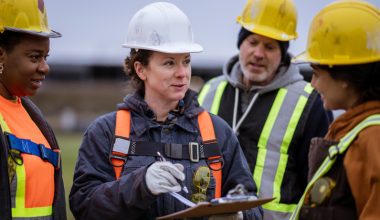Four construction workers in safety gear gathered around a clipboard, discussing plans at a worksite. Faces are blurred for privacy.