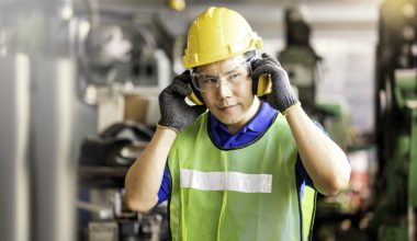 A man wearing a safety vest, hard hat, safety glasses, and protective ear muffs is standing in an industrial plant.