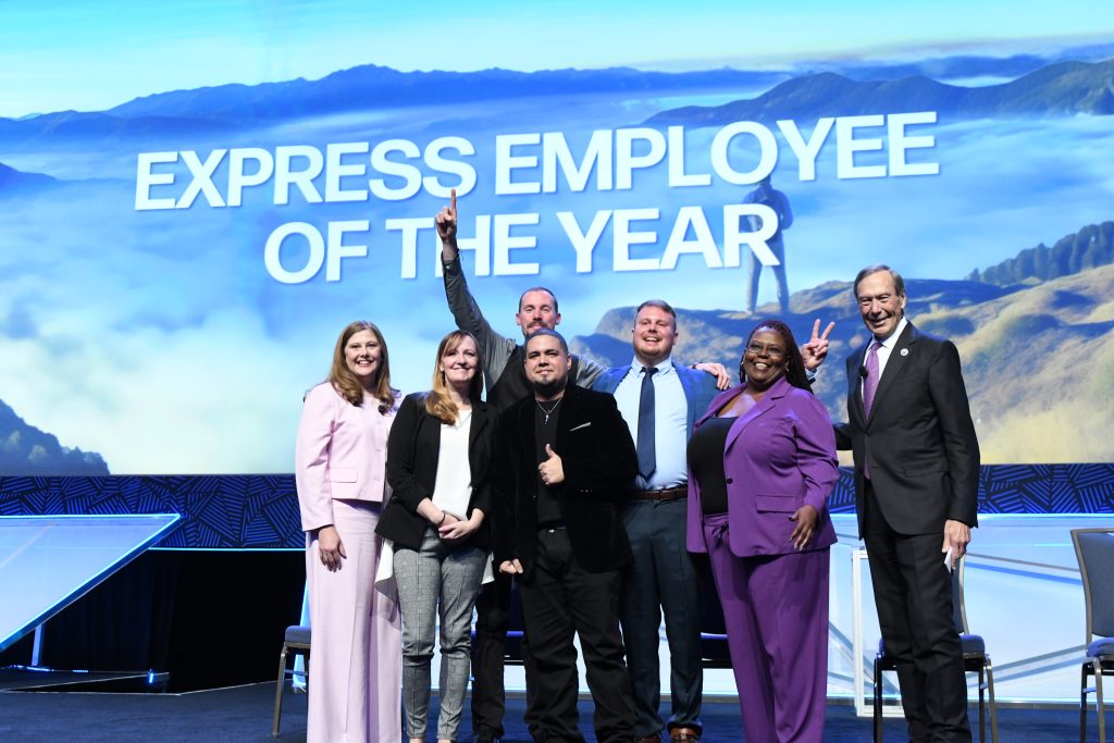 Group of seven people on a stage, one man raising his fist triumphantly, with a backdrop reading "express employee of the year.