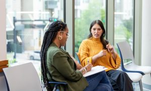 Two women sitting in chairs having a conversation while one takes notes on a clipboard.