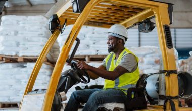 Person operating a yellow forklift in a warehouse, surrounded by stacked pallets wrapped in white plastic, wearing a hard hat and reflective vest.