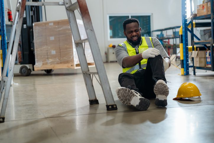 Man in safety gear sitting on warehouse floor, holding his knee in pain next to a yellow hard hat and a step ladder.