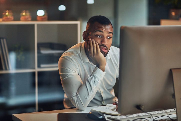 Man in a white shirt sitting at a desk, looking at a computer screen with a bored expression.