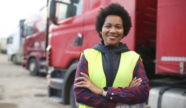 A black woman is wearing a safety vest and standing in front of a row of large parked trucks.