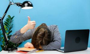 A woman is sitting with her head on her desk beside an open laptop while raising a thumbs up.