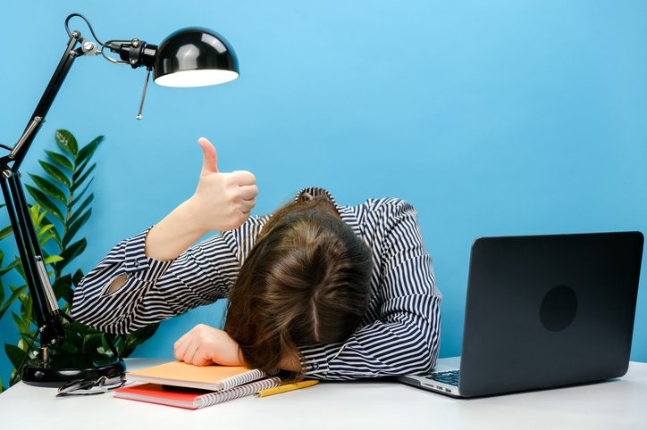 A woman is sitting with her head on her desk beside an open laptop while raising a thumbs up. 