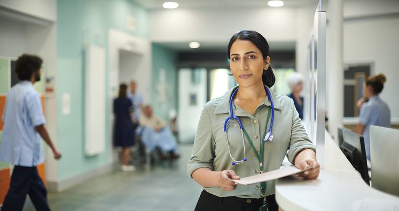 A healthcare professional with a stethoscope stands at a hospital reception desk, holding a clipboard. Behind her, a hospital corridor with staff and a patient in a wheelchair is visible.