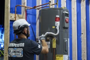 Person in safety gear working on a gray water heater against a blue and wood backdrop.