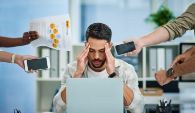 Stressed man at a desk with a laptop, holding their head while multiple hands reach in with phones and documents in an office setting.