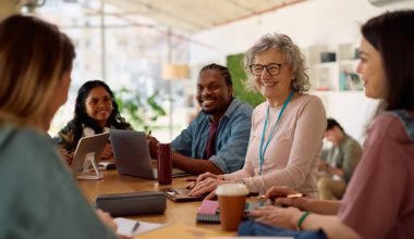 Group of diverse professionals engaged in a collaborative meeting around a table in a modern, well-lit office space. Participants are actively conversing and using laptops and tablets, with notebooks, coffee cups, and office supplies scattered across the table. Large windows provide natural light, enhancing the open and contemporary workspace.