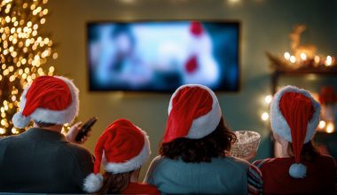 Four people wearing red and white Santa hats sit closely together on a couch, viewed from behind, watching a holiday movie on TV. One holds a remote control, another a bowl of popcorn. A lit Christmas tree and festive lights decorate the cozy room, creating a warm and cheerful holiday atmosphere.