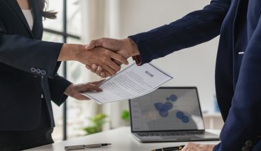 Two people in business attire shaking hands across a desk, one holding a document labeled ‘Resume.’ A laptop displaying a chart and a pen are visible on the desk.