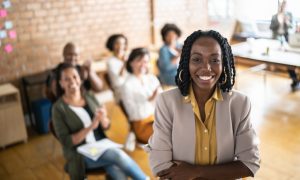 Photo of a Black woman standing in the foreground with arms crossed, wearing a light-colored blazer over a yellow shirt. In the background, several people are seated in a meeting, with a brick wall and sticky notes visible. A table with papers and a laptop is positioned near a window, suggesting a collaborative workspace.
