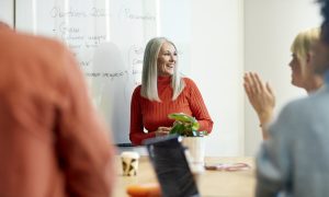Photo of a woman with long gray hair wearing a red turtleneck sweater standing in front of a whiteboard, speaking to two people seated at a table. One person is clapping and another is partially visible in the foreground. Laptops, a mug, and a small plant are on the table.
