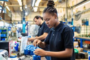 Photo of two assembly line workers in a factory setting; a foreground worker appearing female with hair in a braided bun wears a black short‑sleeve polo shirt and uses a hand tool to assemble blue mechanical components on a workbench, while a background worker appearing male in a black shirt performs a similar task along the same production line.
