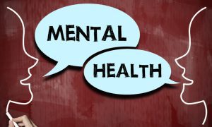 Photo illustration of two outlined human profiles facing each other on a red chalkboard background, with blue speech bubbles reading “MENTAL HEALTH.” A hand holding white chalk appears in the lower-left corner.