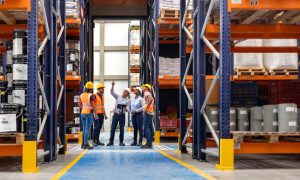 Photo of six adult workers—men and women—wearing yellow hard hats, orange safety vests, long pants, and work boots, standing in a warehouse aisle between tall pallet racks filled with boxed and bagged inventory; one person gestures upward while others look on, suggesting a team discussion or inspection.