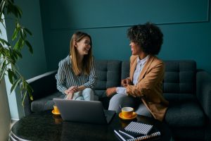 Two young businesswomen are networking in a quiet coffee shop, collaborating over a laptop in a calm, introvert‑friendly setting that supports building referrals without small talk.