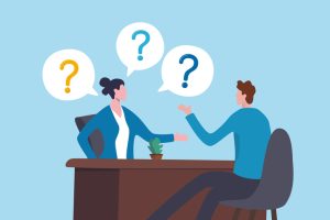Illustration of two office workers seated across a desk in conversation, with speech bubbles containing question marks above them, symbolizing a discussion, interview, or Q&A session; blue background, brown desk, and small potted plant on the tabletop.